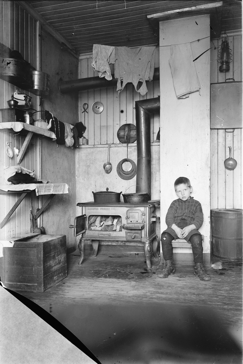 Simen Baardseth, INTERIØR, ARBEIDERHJEM, HUSMANNSPLASS, GUTT SITTER PÅ KJØKKEN MED VEDKOMFYR fra Moelven Støberi.