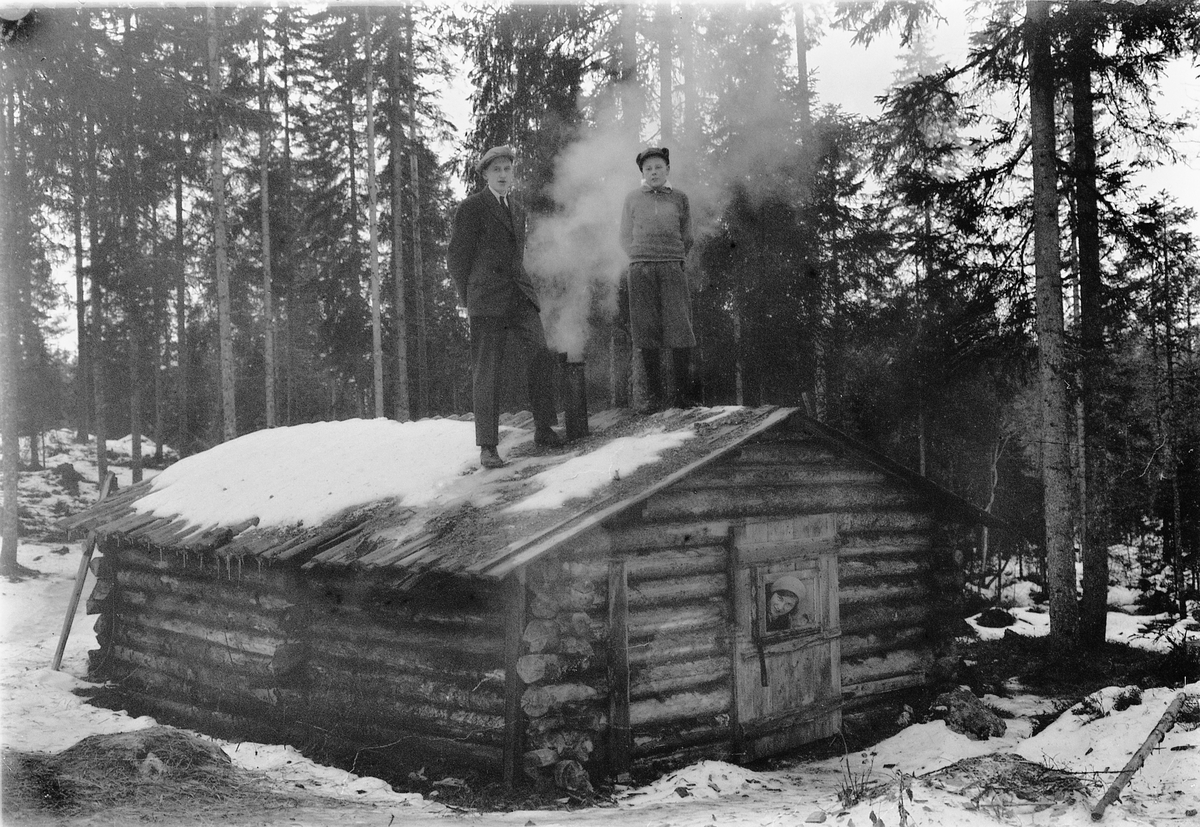Simen Baardseth, Tømmerkoie i skogen. Sverre S. (sønn) på taket, ukjent kvinne stikker hodet ut av døra.