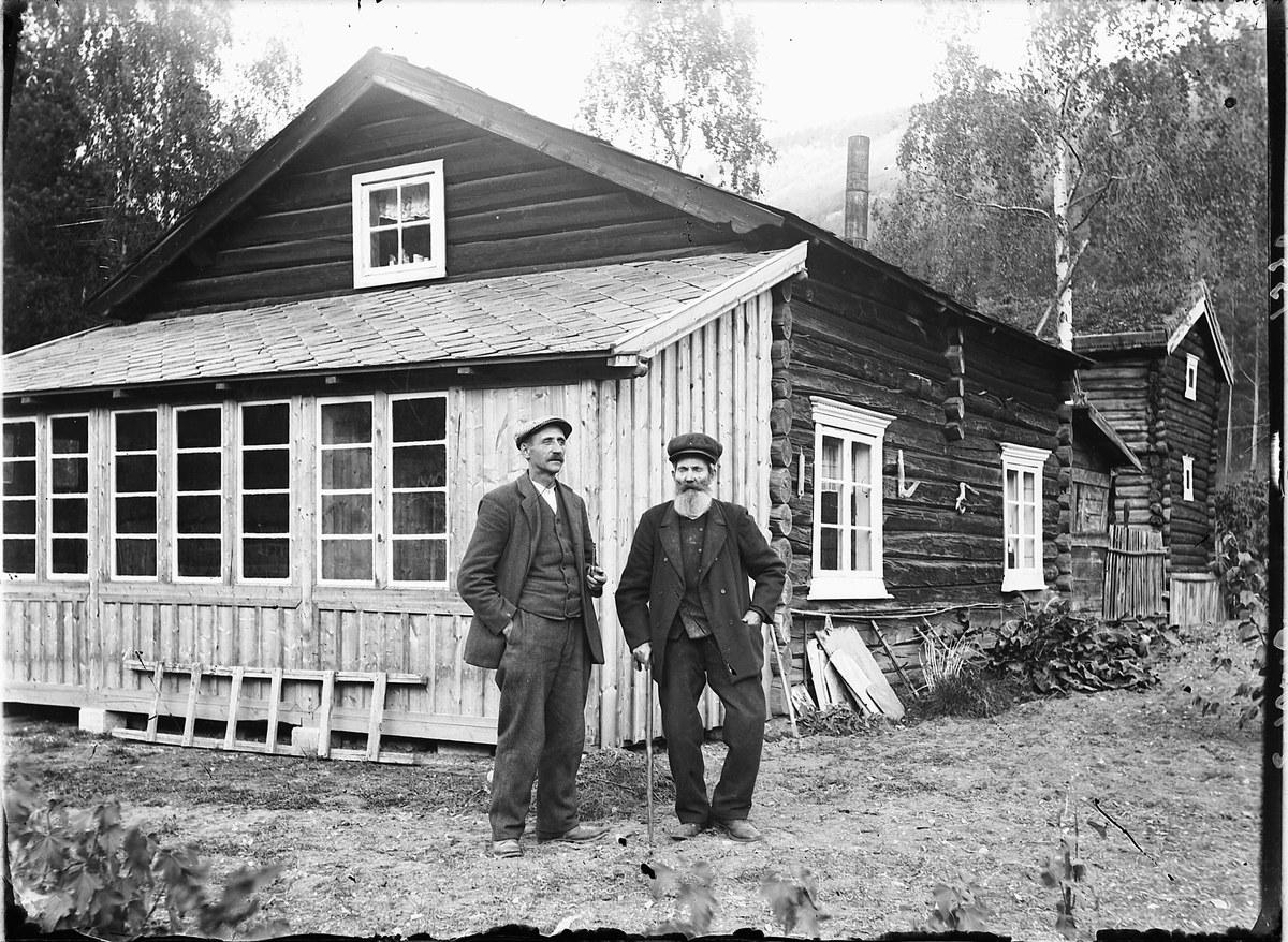 Simen Baardseth, To eldre menn foran et tømmerhus.