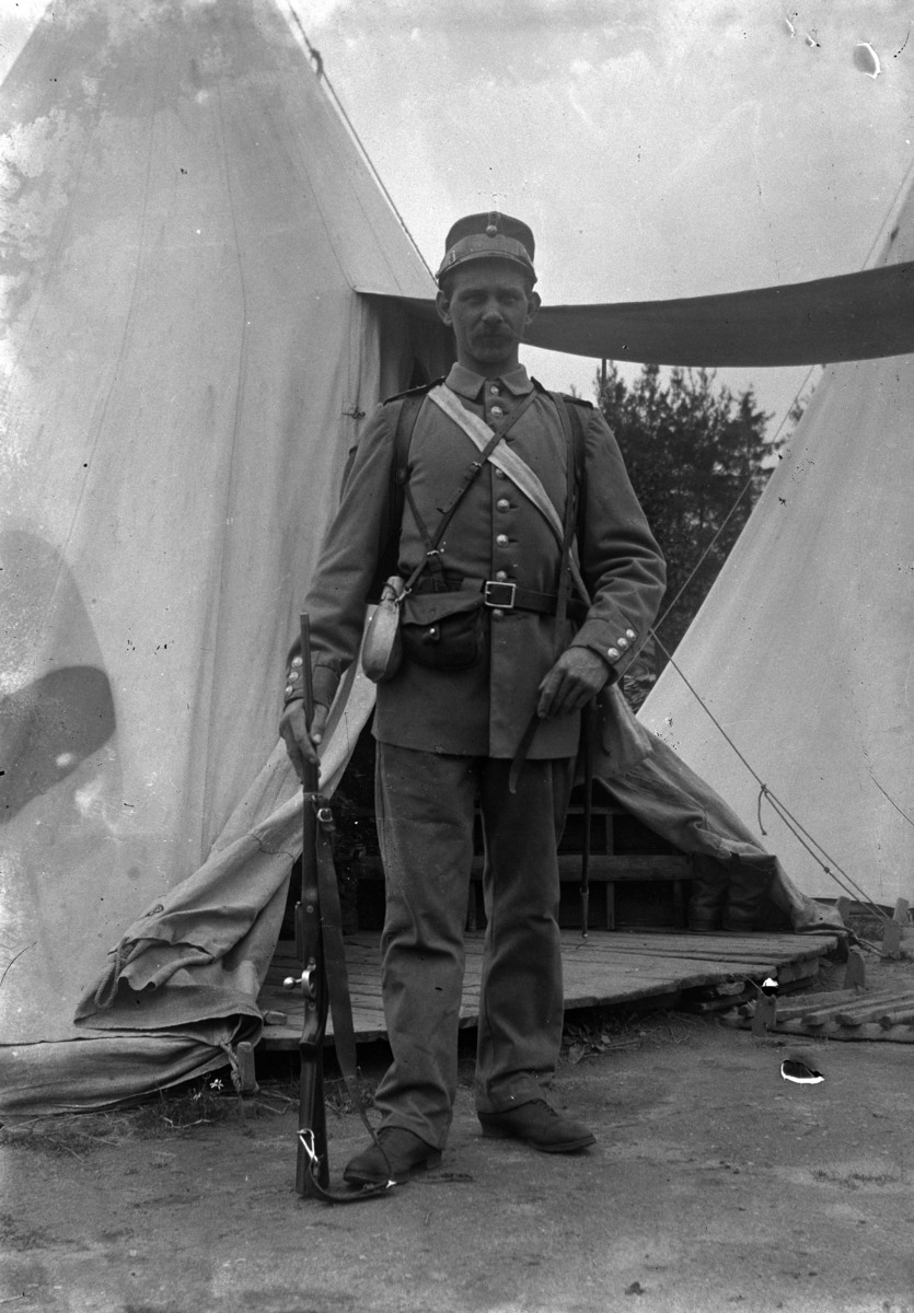 Johannes Larsen Alhaug, En soldat med uniform og våpen foran et telt.