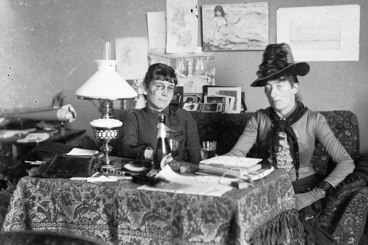Hans Emil Erichsen, 2 kvinner ved bord. Interiør stue. Kvinnen til venstre er Vincentia Sofie Emma Guidotti Skramstad (1852 - 1944).