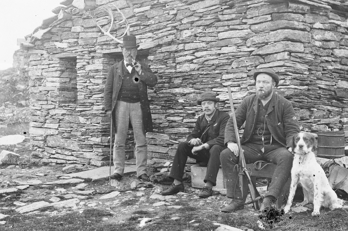 Hans Emil Erichsen, 3 menn med hund og gevær ved Friisbua på Ringebufjellet. Fra venstre er J.A. Friis, Ludvig Skramstad, ukjent. Denne steinhytta ble bygget av forfatteren og professoren J. A. Friis i 1878. Friis ferierte her i fjellet i mange år, noe han har beskrevet i boka ”Til fjeld i Ferierne. Jæger og Fiskerliv…