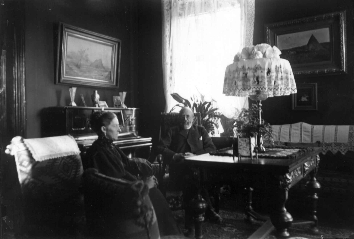 Hermann Christian Neupert, Interiør, ukjent sted. 1908-1910. Stue. Kvinne og mann ved bordet. Malerier. Piano.