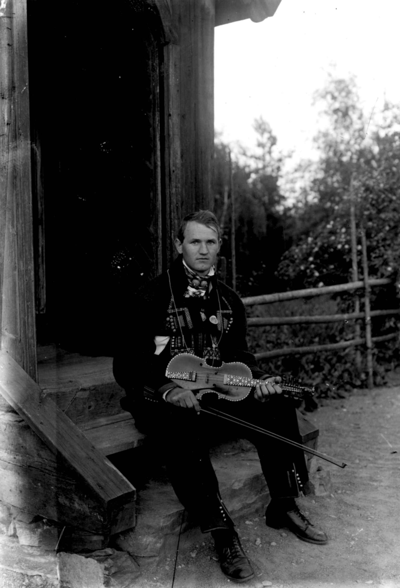 Hermann Christian Neupert, Mannsdrakt, Setesdal, Aust-Agder. 1925. Spelemann med hardingfele kledd i Setesdalsbunad. Sittende på en trapp.
