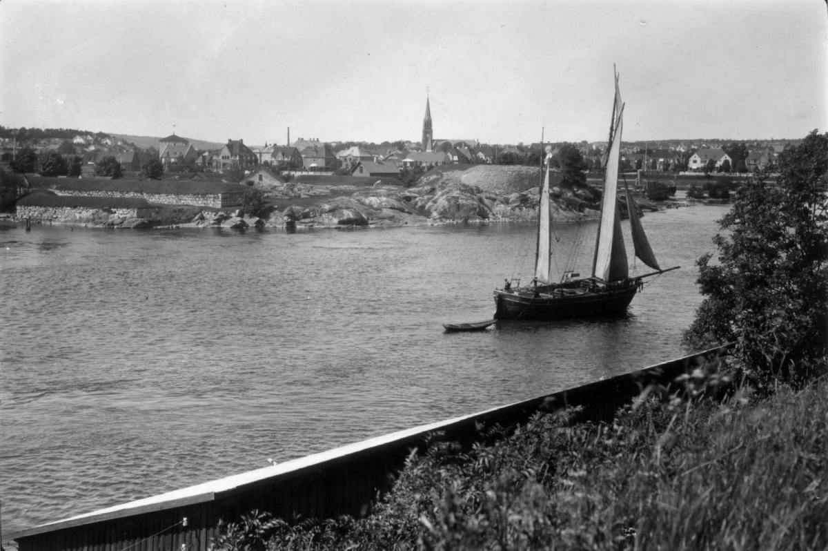 Hermann Christian Neupert, Fredrikstad, Østfold. 1930. Oversiktsbilde med elv og seilskute i forgrunnen. Bebyggelse med kirke i bakgrunnen.