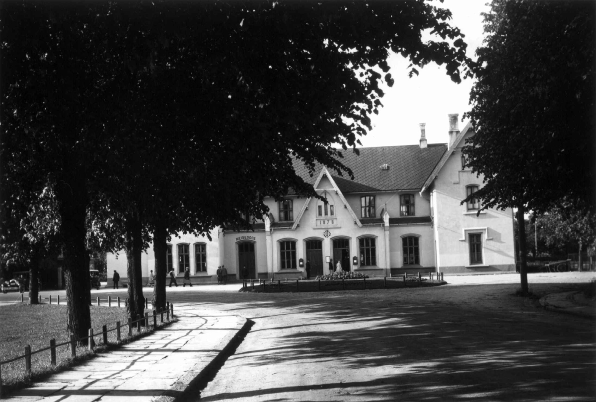 Hermann Christian Neupert, Fredrikstad. 1930. Gatemotiv. Allé med trær som fører til jerbanestasjonen (?), en murbygning fra 1878. Over en inngang er det skrevet reisegods. Mennesker på vei inn i bygningen.