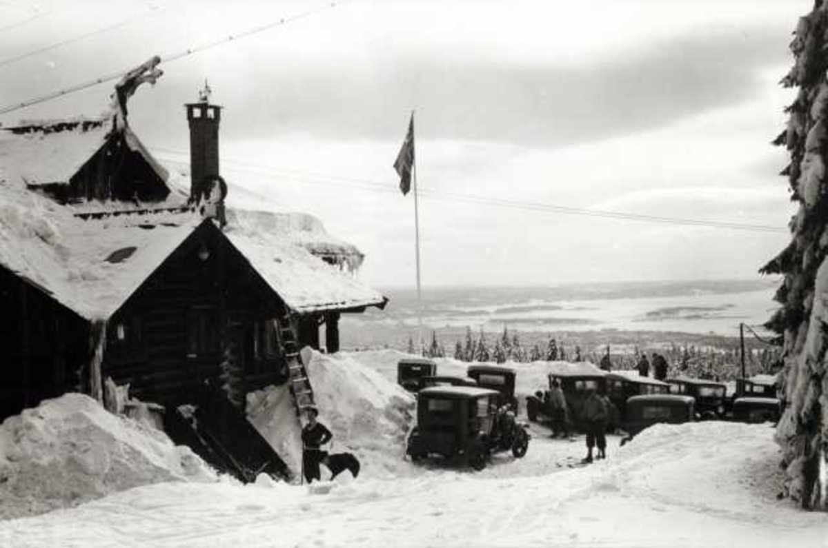 Hermann Christian Neupert, Frognerseteren Restaurant, Nordmarka, Oslo. Vintermotiv. Utsikt over byen. Mennesker og biler foran restauranten. Flaggstang med flagg.