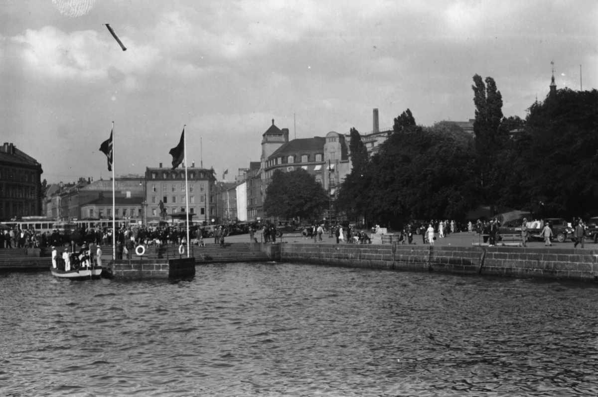 Hermann Christian Neupert, Oslo havn. Brygge med flagg, båt med gaster og fotgjengere langs kaia. Bilde tatt fra sjøen.