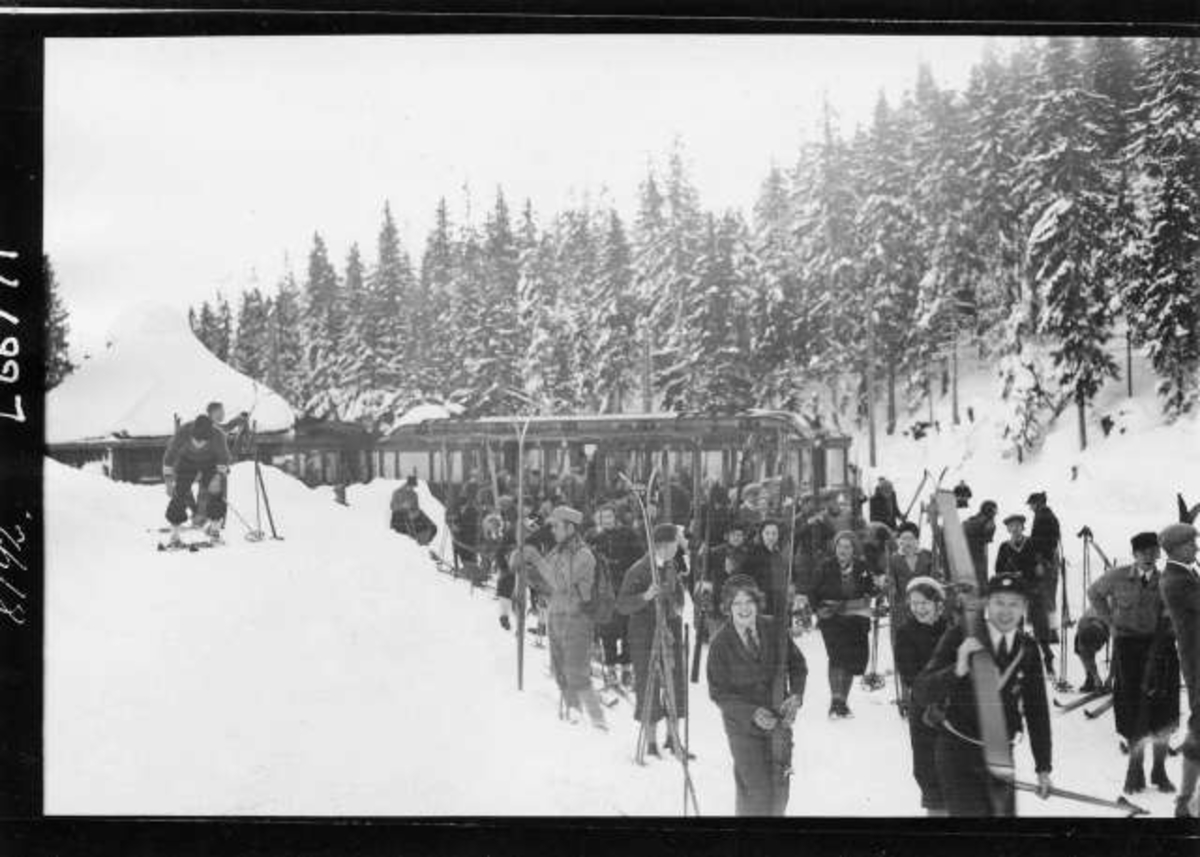 Hermann Christian Neupert, Vintermotiv. Stasjon på Holmenkollbanen med skog i bakgrunnenn. Trikk på plattformen. Skiløpere på vei ut i løypa.