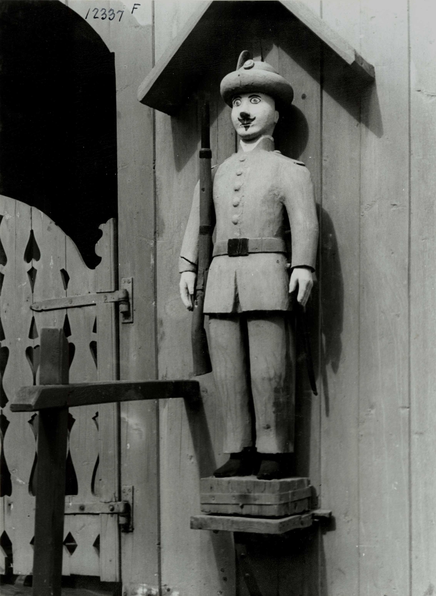 Anders Beer Wilse, Lensmannsgården Østgård, Tolga, Hedmark ant. ca. 1940. Detalj av stabbur med trefigurer på fronten, figurene skal være laget av fanger. Soldat/jeger.