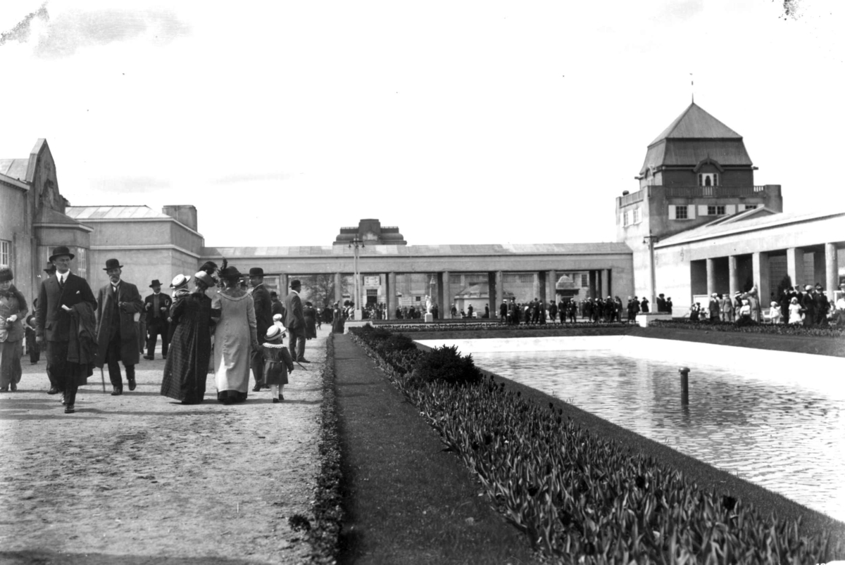 Hermann Christian Neupert, Jubileumsutstillingen på Frogner, Oslo, 1914. Frogner Hovedgård med Bergen bys paviljong til venstre. Publikum spaserer på området. Basseng til høyre. Blomsterbed langs kanten.