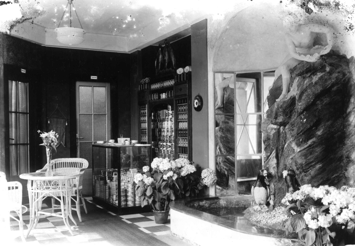 Hermann Christian Neupert, Jubileumsutstillingen på Frogner, Oslo, 1914. Interiør paviljonger. Skulptur til høyre i bildet. Blomster. Reklameprodukter i hyller i bakgrunnen.