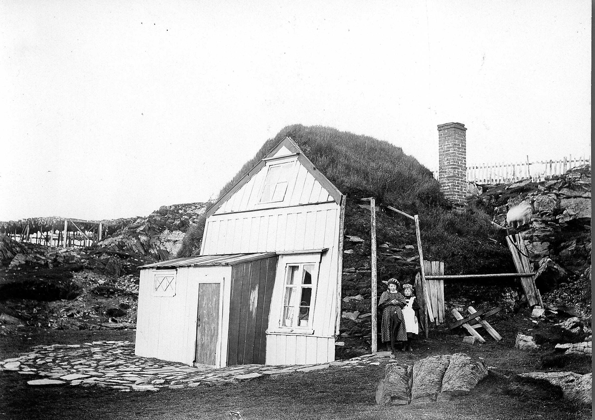 Robert Collett, Bolighus, ukjent sted, med panelt gavlside med steinvegger bygget inn mot fjellvegg. To jenter står utenfor, den ene med en paraply i hånda. Serie tatt av Robert Collett (1842-1913), amatørfotograf og professor i zoologi.