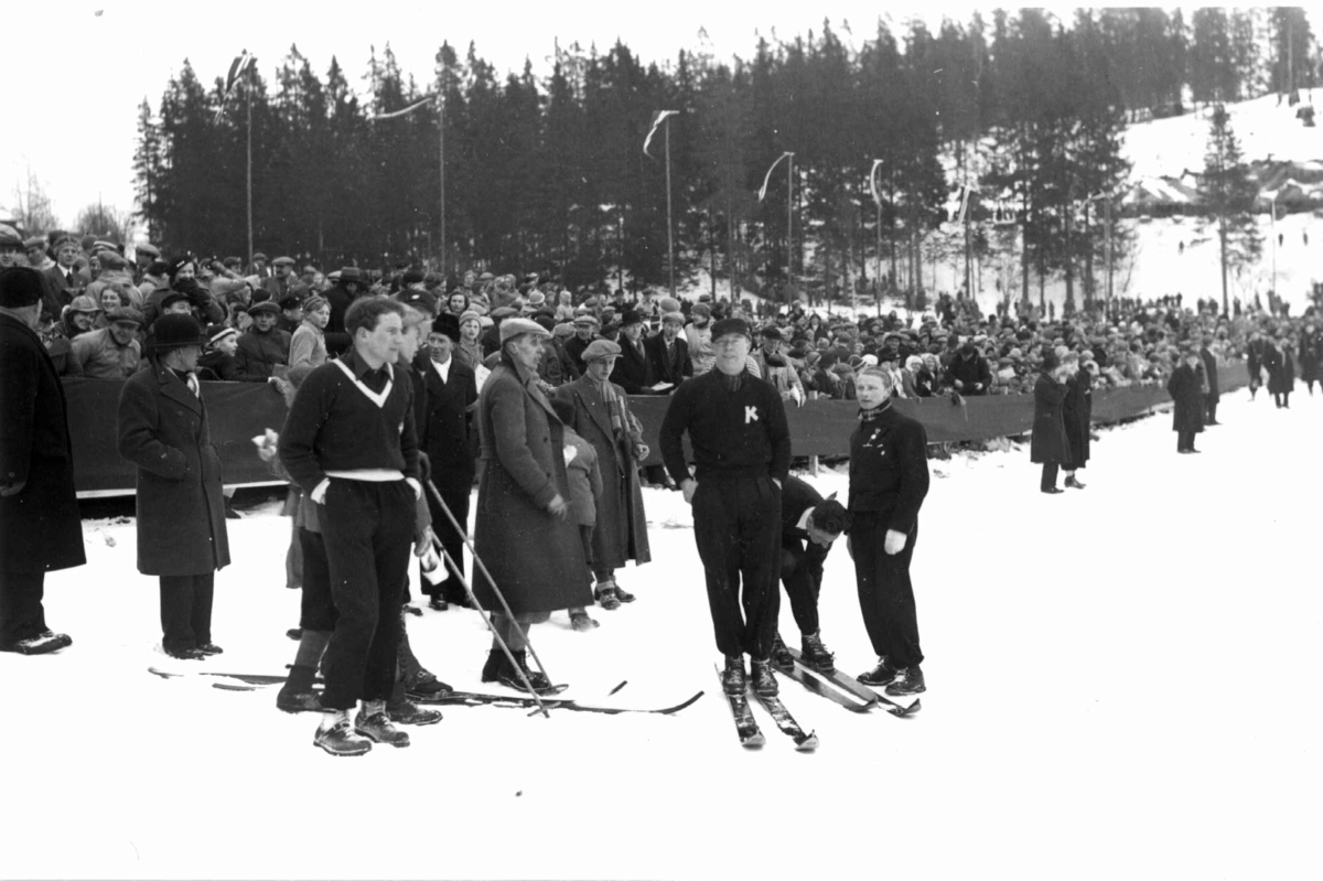 Hermann Christian Neupert, Ski. Hopprenn. Holmenkollen, Oslo 1934. Hoppere og funksjonærer står på sletta. Publikum i bakgrunnen.