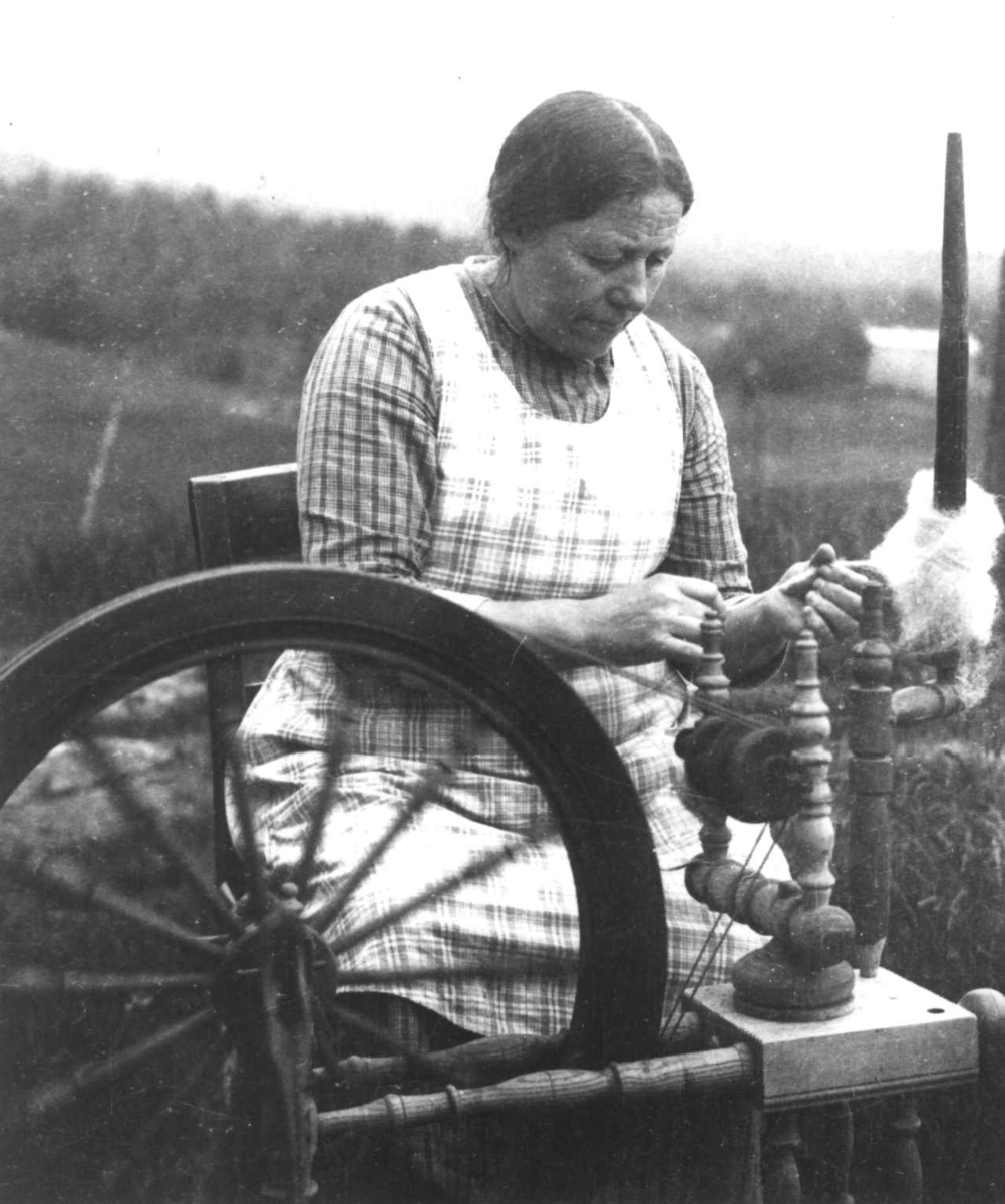 Anna Grostøl, En kvinne sitter ved en rokk og spinner. Ukjent sted 1935.