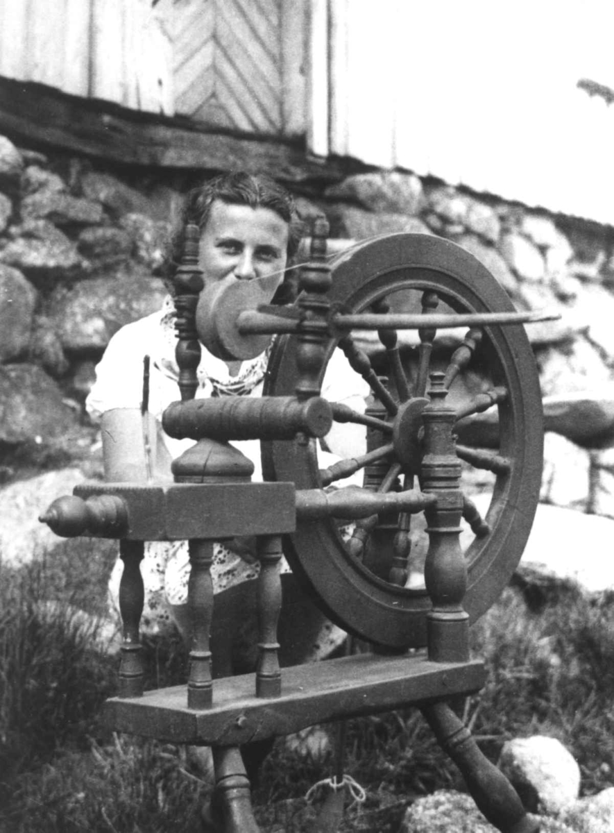 Anna Grostøl, En kvinne sitter ved rokken og spinner. Ukjent sted 1935.
