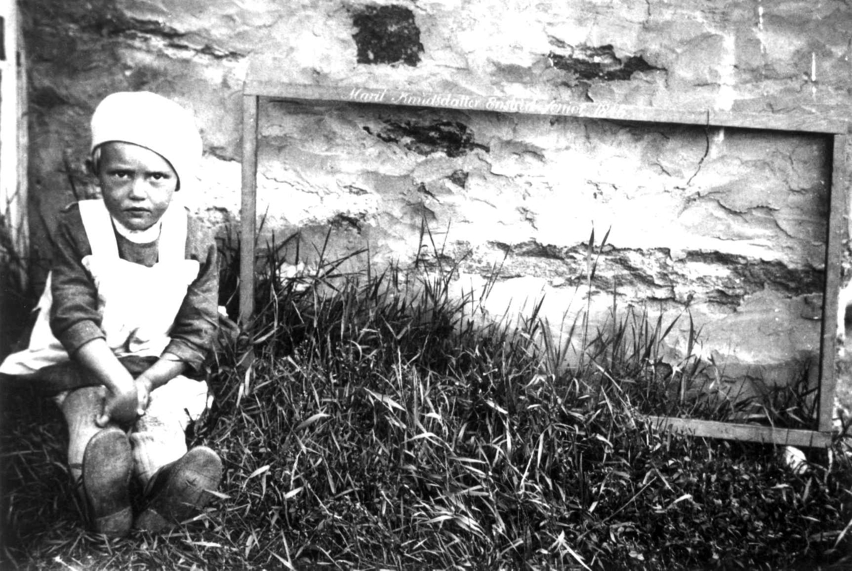 Anna Grostøl, Antagelig Lesja, Oppland 1934. Liten jente sitter ved siden av en sprangramme ved en husvegg.