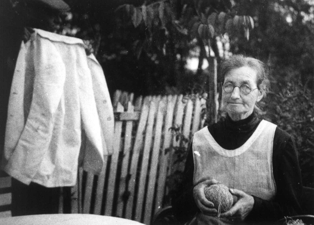Anna Grostøl, Portrett av Synnøve Kvale som sitter i hagen. Ved siden av henne står kunstmaler Müller som viser frem en jakke. Tveit, Kristiansand 1937.
