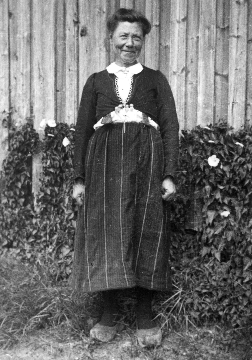 Anna Grostøl, Fru Tarjgjerd Hellesvik står utenfor en husvegg. Eiken, Hægesbostad, Vest-Agder 1937.