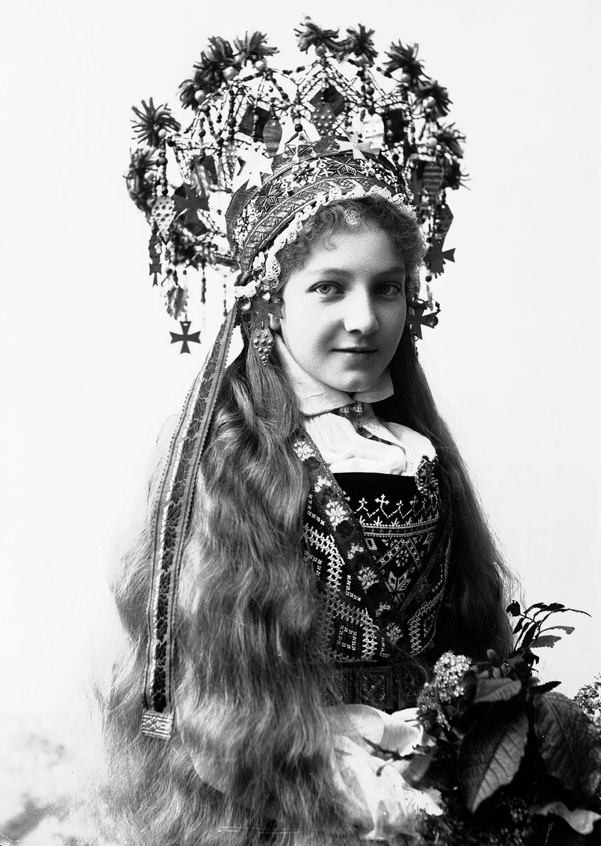 Solveig Lund, Studiofotografi av kvinne med brudedrakt og brudekrone. Sittende med blomster i fanget. Utslått hår. Hofteutsnitt. Vendt mot høyre. Ser på betrakteren.