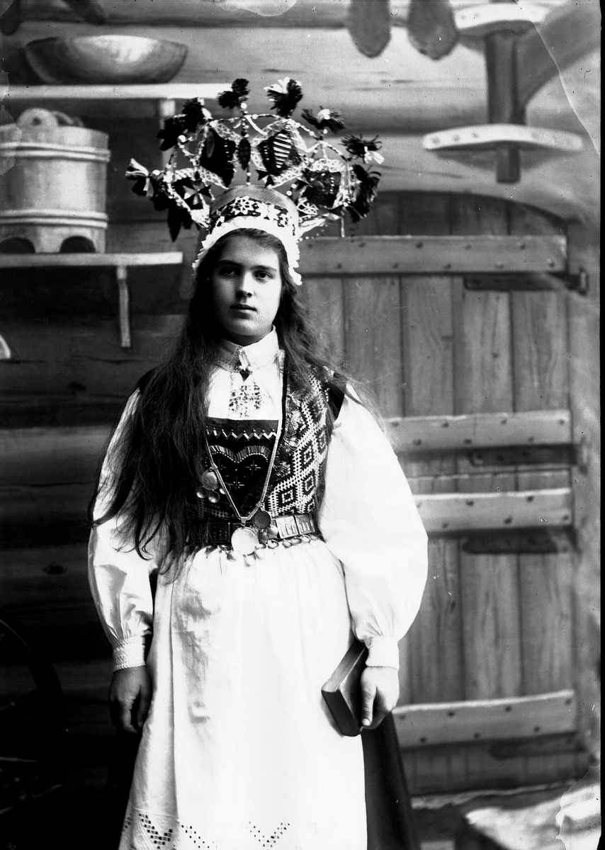 Solveig Lund, Studiofotografi av kvinne med brudedrakt og brudekrone stående foran malt bakgrunn. Denne viser interiør fra seter - vegger, dør, boller og tine. Holder salmebok/bibel i hånden. Utslått hår. 3/4 dels utsnitt. Frontalt vendt. Ser ut av bildet.