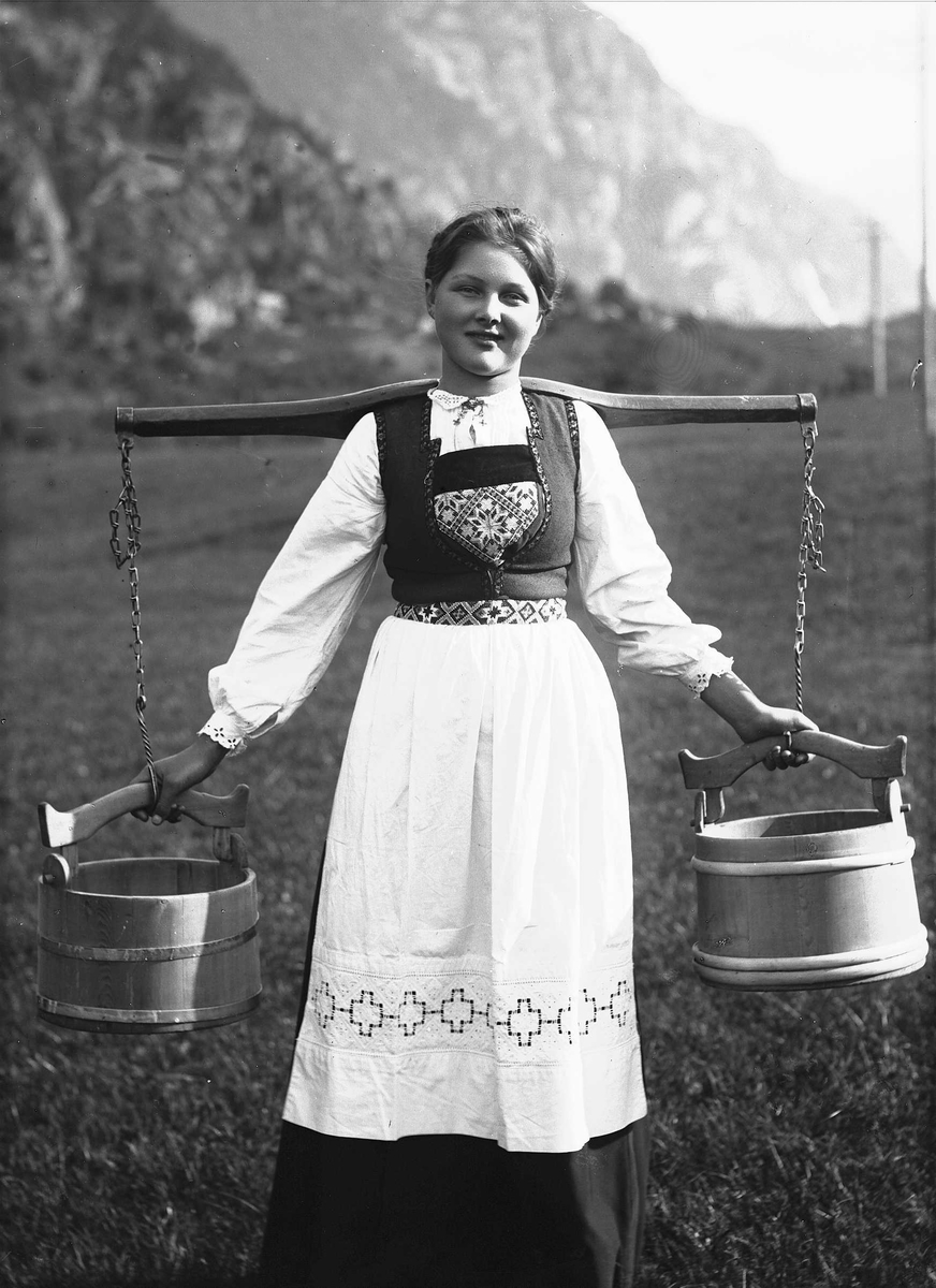 Solveig Lund, Kvinne i Hardangerdrakt står på tun, bærer på to vannbøtter. I bakgrunnen sees fjell.