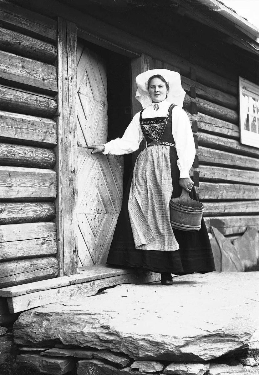 Solveig Lund, Kvinne i Hardangerdrakt står i inngangsdøren til seter. Holder en bøtte i hånden.