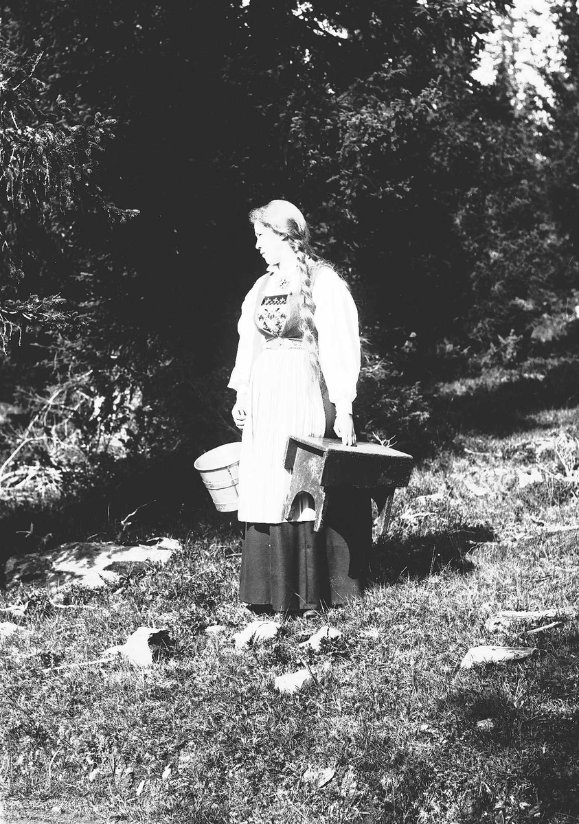 Solveig Lund, Kvinne i drakt stående på jorde med bøtte og krakk i hånden.