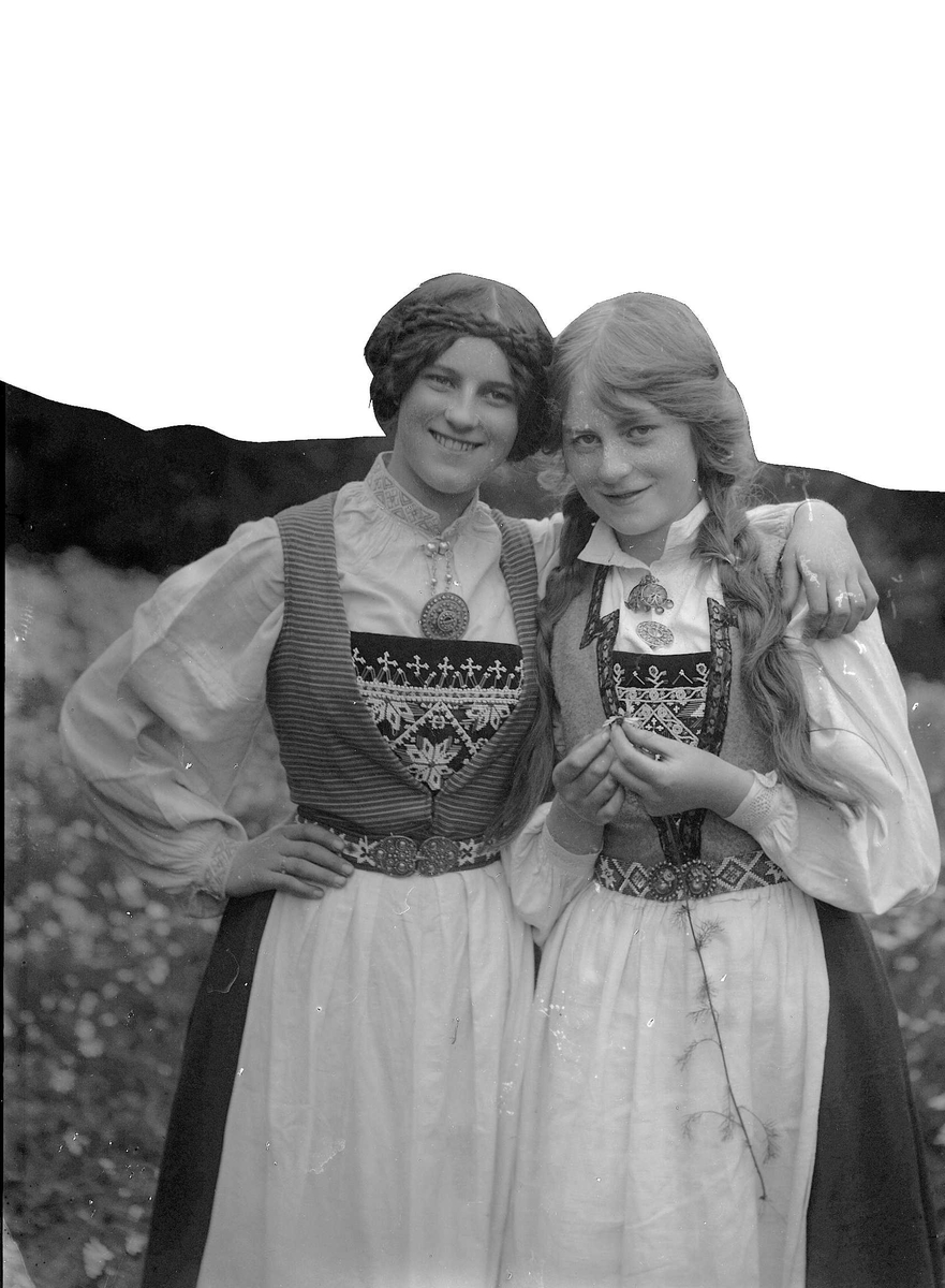 Solveig Lund, To kvinner i nasjonaldrakt står med armene rundt hverandre på blomstereng. Den ene holder en prestekrage i hendene.