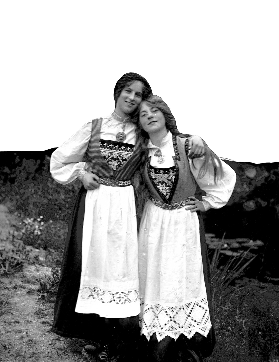 Solveig Lund, To kvinner i nasjonaldrakt står med armene rundt hverandre. Vann i bakgrunnen.