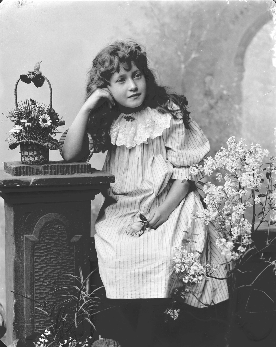 Solveig Lund, Portrett av jente med kjole. Hun sitter og støtter hodet med den ene hånden på en stolpe og er omgitt av blomster. Kikker ut mot høyre i bildet. Bakgrunnen er malt med trær og arkitekturelementer.