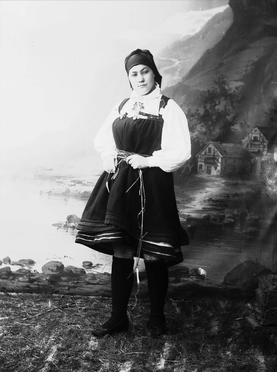Solveig Lund, Studioportrett av kvinne i nasjonaldrakt . Hun holder strå i hendene og kikker ned mot gulvet. Står på gress/jordbunn. Malt bakgrunn med fjord og fjell og et lite hus.