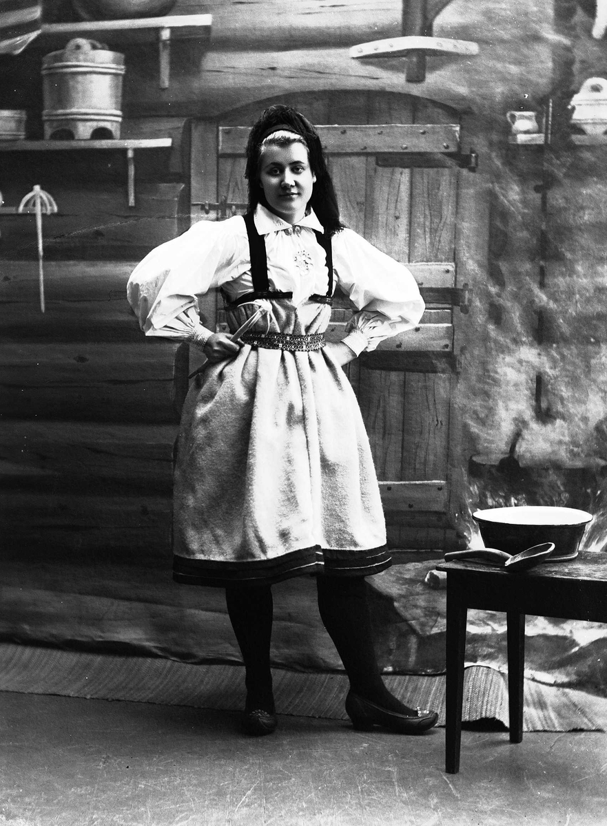 Solveig Lund, Studioportrett av kvinne i nasjonaldrakt. Hun står med armene på hoften og holder en sleiv i den ene hånden. Ved siden av står et bord med bolle og sleiv. Kvinnen ser ut mot venstre. Bakgrunnen er malt og viser seterinteriør. Til høyre står kjelen på ildstedet og til venstre står vegghylle med tine…