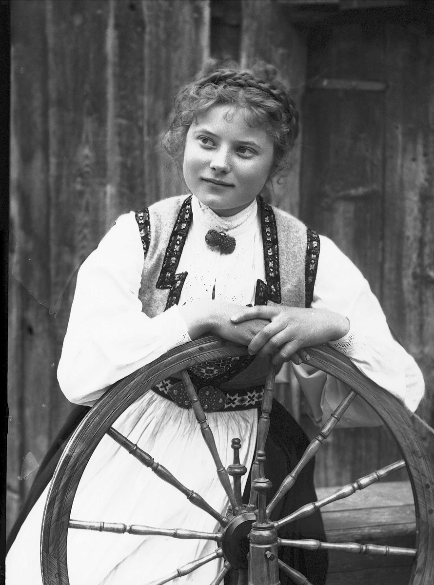 Solveig Lund, Studioportrett av kvinne i nasjonaldrakt som lener seg mot hjulet på en rokk. Kvinnen ser mot vensre i bildet. Bakgrunnen er malt og viser trevegger.