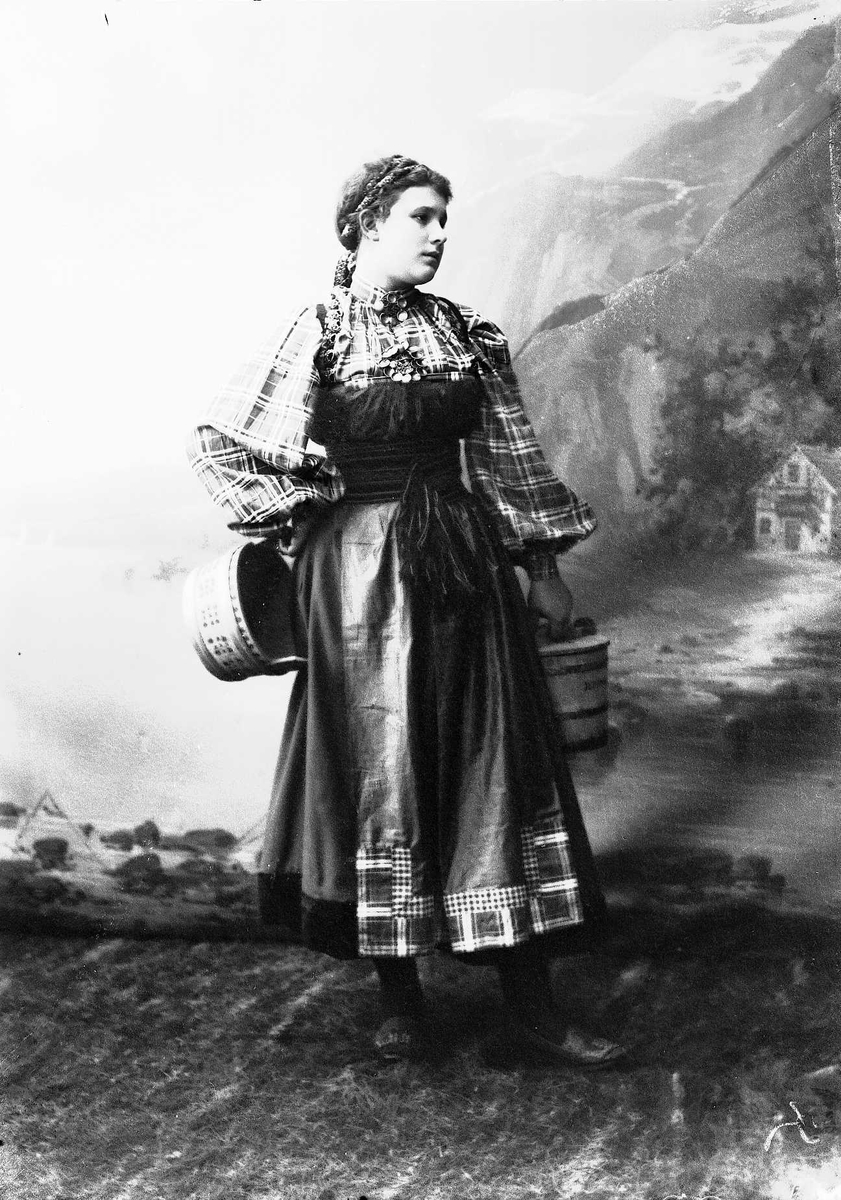 Solveig Lund, Studioportrett av kvinne i beltestakk som holder to tiner.Står på gress. Bakgrunnen er malt med fjord og fjell landskap. Kvinnen har korsett, påkledd som nasjonalromantisk motiv.