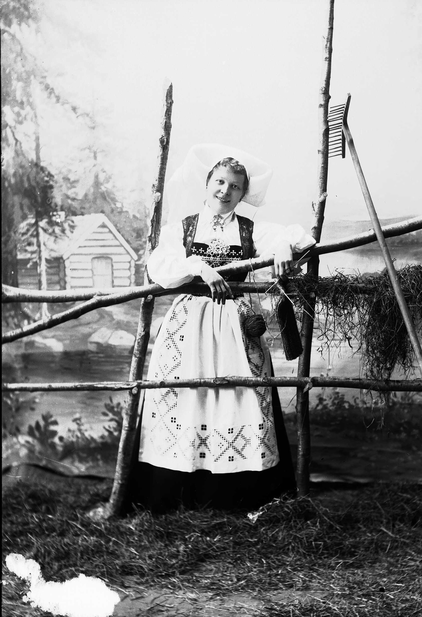 Solveig Lund, Studioportrett av kvinne i nasjonaldrakt. Hun lener seg mot hesjegjerde og holder strikketøy i hendene. Bakgrunnen er malt med skog, vann og trehytte, på gulvet er det høy/gress. Rive er lent mot gjerdet.