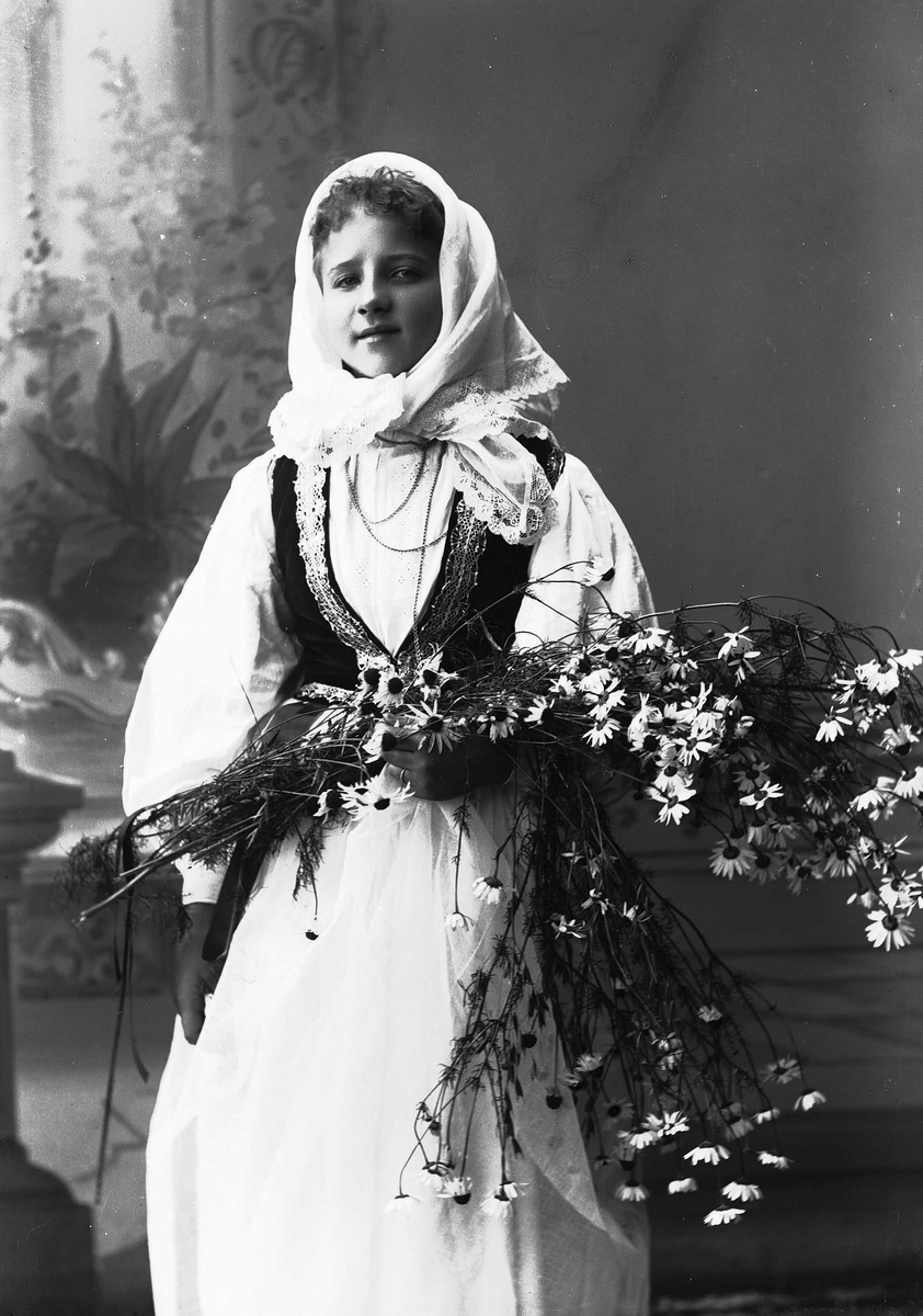 Solveig Lund, Studioportrett av kvinne i drakt med skaut. Holder blomster i hendene. Bakgrunnen er malt med blomster og arkitekturelementer.