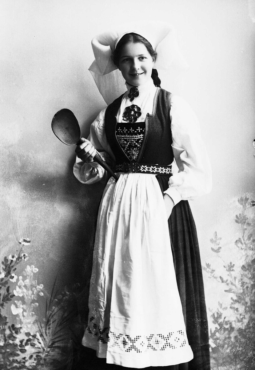 Solveig Lund, Studioportrett av kvinne i drakt med hodeplagg. Holder sleiv i den ene hånden. Bakgrunnen er malt med blomster.