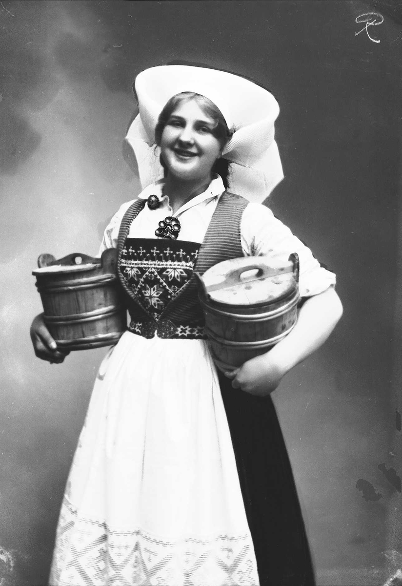 Solveig Lund, 3/4 portrett av kvinne i drakt med skaut, tatt i fotografens studio. Hun holder en tine under hver arm. Nøytral bakgrunn.