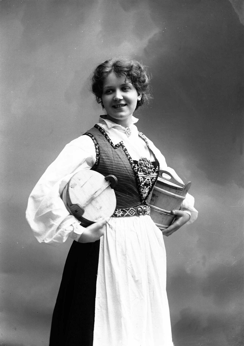 Solveig Lund, 3/4 portrett av kvinne i drakt, tatt i fotografens studio. Hun holder en tine under hver arm. Nøytral bakgrunn.