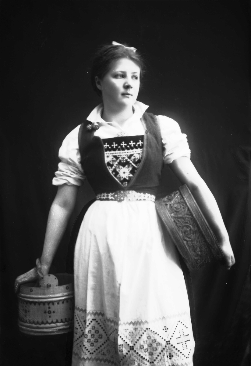 Solveig Lund, 3/4 portrett av kvinne i drakt, tatt i fotografens studio. Hun holder en bøtte i høyre hånd og en eske under venstre arm. Nøytral bakgrunn.