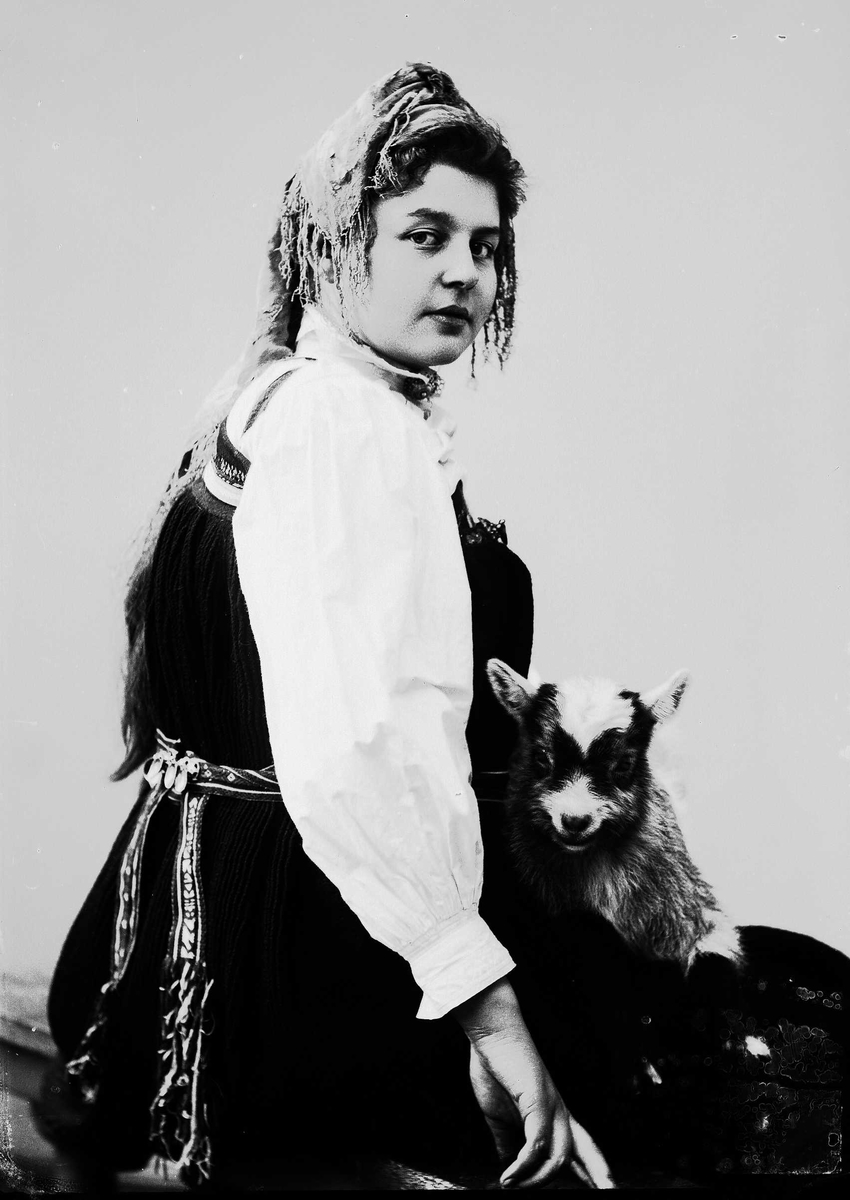 Solveig Lund, 1/2 portrett av kvinne i drakt og skaut, tatt i fotografens studio. Hun sitter med høyre side vendt mot fotografen og kikker ut på betrakteren. Hun holder en geit på fanget. Nøytral bakgrunn.