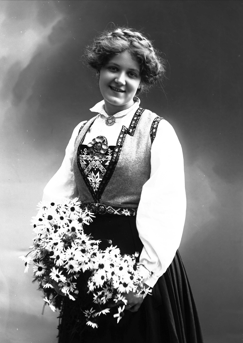 Solveig Lund, 3/4 dels studioportrett av kvinne i drakt og oppsatt hår. I hendene holder hun en favn med prestekrager. Bakgrunnen er malt med himmel og skyer.