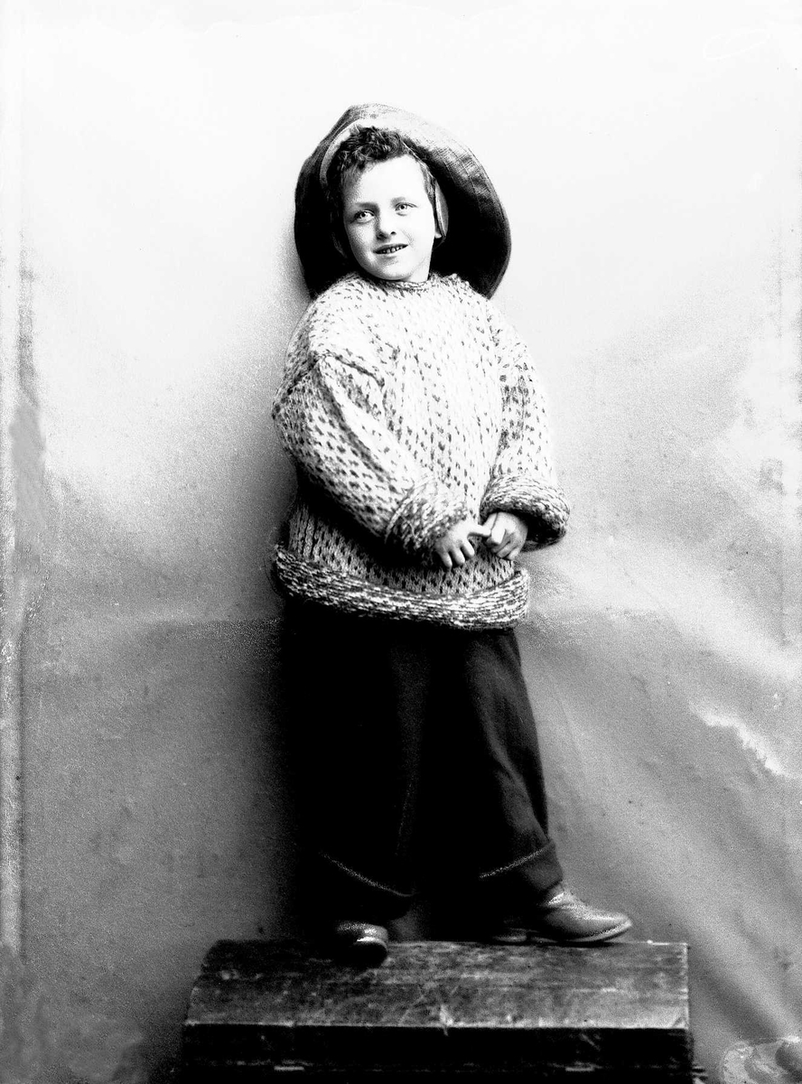 Solveig Lund, Studioportrett av gutt i drakt og sydvest. Han står på en benk og er avfotografert mot en nøytral bakgrunn.