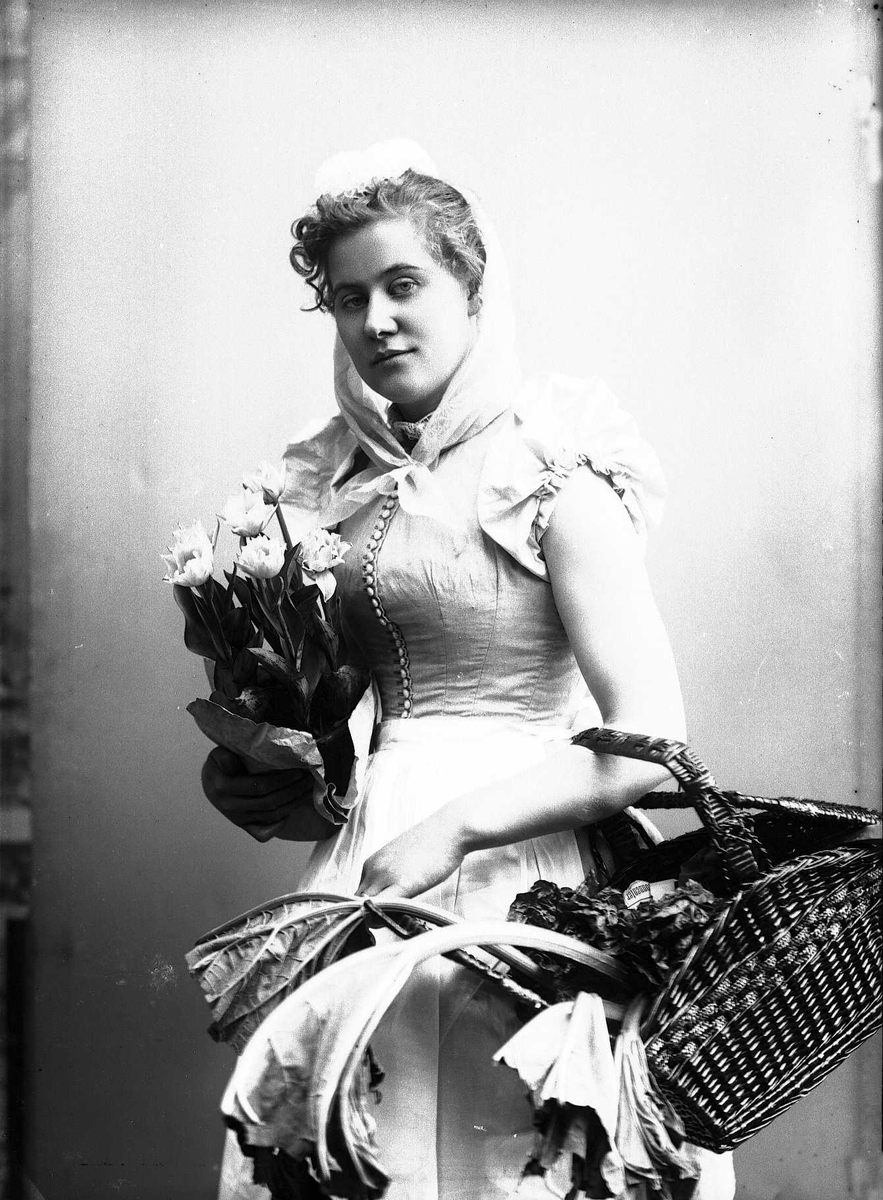 Solveig Lund, 3/4 dels studioportrett av kvinne i drakt og skaut. I høyre hånd holder hun en bukett blomster. I venstre en kurv med grønnsaker. Avfotografert mot en nøytral bakgrunn.