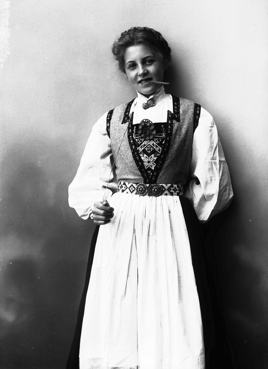 Solveig Lund, 3/4 dels studioportrett av kvinne i drakt, med oppsatt hår. Hun biter på et strå hun holder i høyre hånd. Hun er avfotografert mot en nøytral bakgrunn.