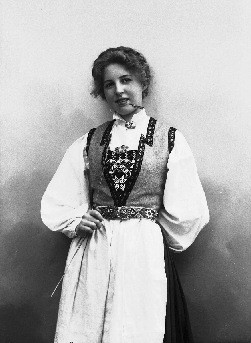 Solveig Lund, 3/4 dels studioportrett av kvinne i drakt, med oppsatt hår. Hun biter på et strå hun holder i høyre hånd. Hun er avfotografert mot en nøytral bakgrunn.