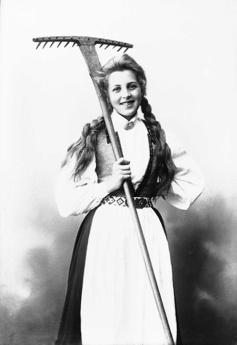 Solveig Lund, 3/4 dels studioportrett av kvinne i drakt, med flettet hår. Hun holder en rive i høyre arm, mens den venstre hviler mot hoften. Bakgrunnen er nøytral.