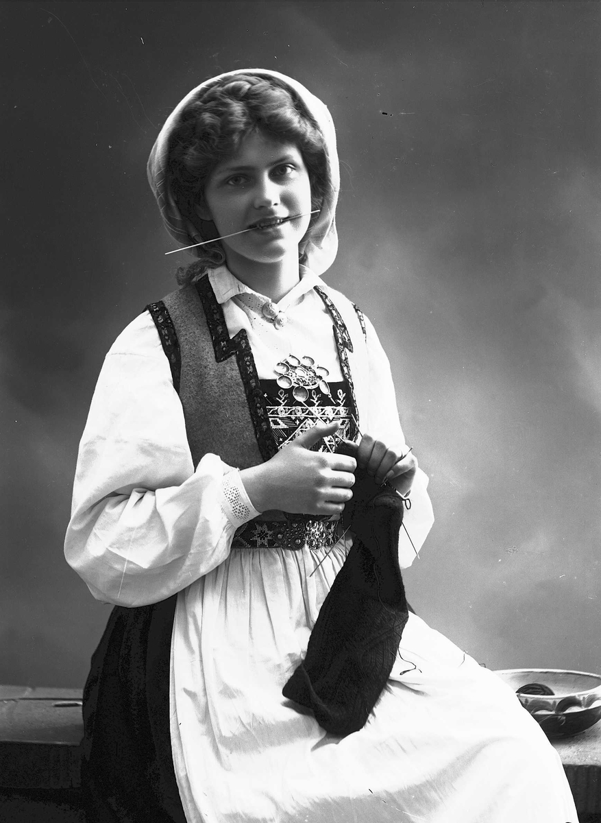 Solveig Lund, 1/2 dels studioportrett av kvinne i drakt og skaut, som sitter på en benk og strikker. Til høyre for henne står en bolle. Kvinnen holder en strikkepinne i munnen. Bakgrunnen er nøytral.