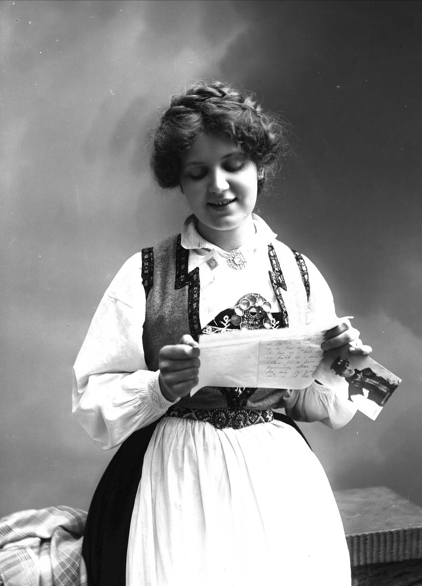 Solveig Lund, 1/2 dels studioportrett av kvinne i drakt, som sitter på en benk og leser et brev. I venstre hånd holder hun et fotografi av en mann. På benken til høyre for henne ligger et sjal. Bakgrunnen er nøytral.